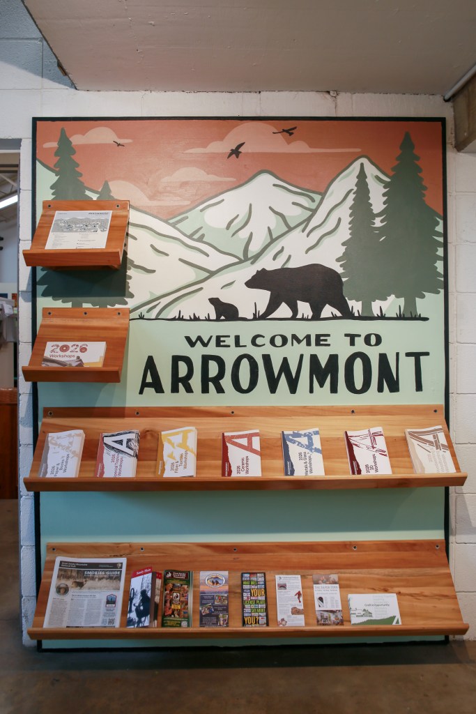 A functional art installation by Annie Fox Design at the Arrowmont School of Arts and Crafts. The central feature is a custom-painted mural of the Great Smoky Mountains, designed in a vintage screen-printed poster style. It depicts a mother bear and cub, geometric pine trees, and hand-lettering that reads "WELCOME TO ARROWMONT." The mural is integrated with multiple floating and tiered wood shelves, which are filled with 2026 workshop catalogs, guides, and brochures, illustrating how design and utility intersect in a physical space.