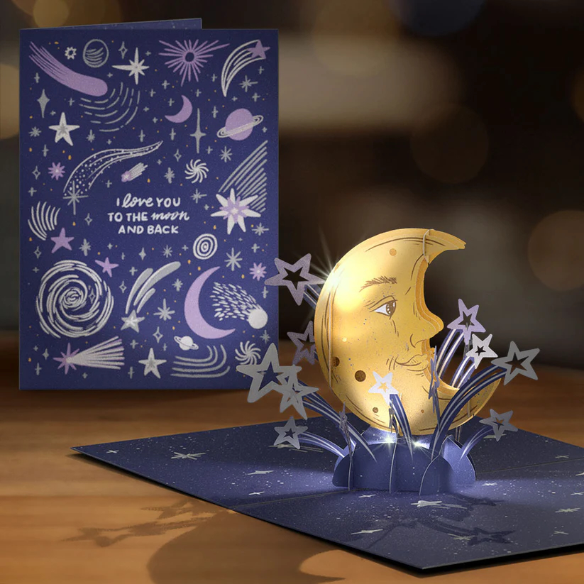 A celestial-themed greeting card illustration for Lovepop by freelance designer Annie Fox Design. The design features a navy blue background filled with lavender and white hand-drawn stars, comets, planets, and galaxies. In the center, the phrase "I love you TO THE moon AND BACK" is written in a combination of custom sans-serif and script lettering. The artwork features a soft, chalky digital texture optimized for art licensing.
